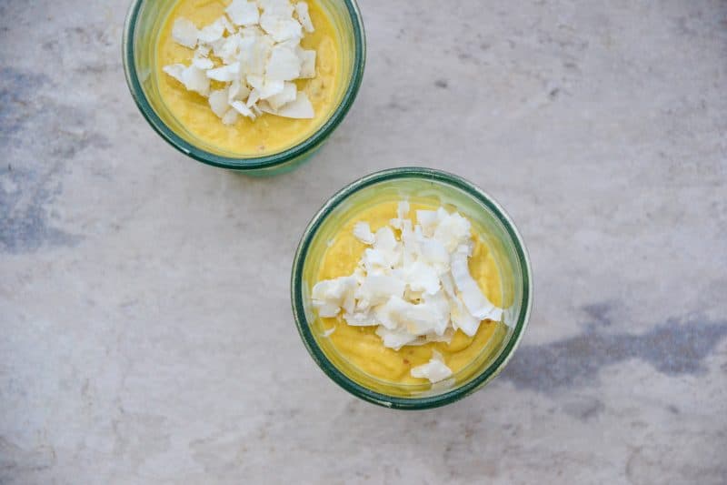 5 ingredient mango pudding recipe with coconut flakes on top and creamy pudding inside two glasses for dessert on a marble table.